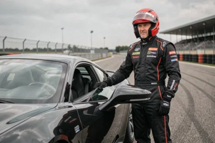 Pilote en combinaison de course rouge posant à côté d'une voiture sportive noire dans le paddock d'un circuit de pilotage